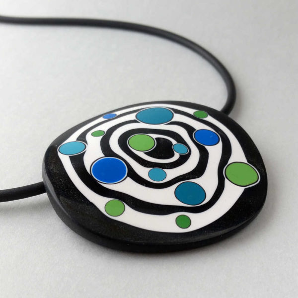 Large handmade pendant with organically-shaped concentric black and white circles, and irregular dots of bright blue and green. It is approximately 6.6cm in diameter and hangs on a black adjustable cord.