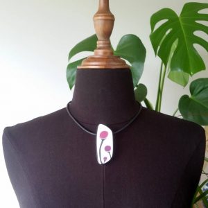 Handmade shield shaped necklace featuring an abstract flower bud motif in plum on a white background, with charcoal border. It is approximately 2.6 cm wide and 6.4cm long and hangs on a black adjustable cord.