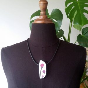 Handmade shield shaped necklace featuring an abstract flower bud motif in plum on a white background, with charcoal border. It is approximately 2.6 cm wide and 6.4cm long and hangs on a black adjustable cord.