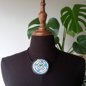 Large handmade pendant with organically-shaped concentric black and white circles, and irregular dots of bright blue and green. It is approximately 6.6cm in diameter and hangs on a black adjustable cord.