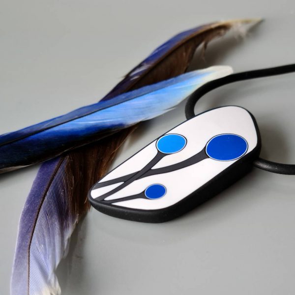 Handmade shield shaped necklace featuring an abstract flower bud motif in blue on a white background, with charcoal border. It is approximately 2.6 cm wide and 6.4cm long and hangs on a black adjustable cord.